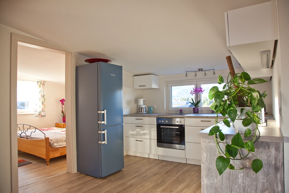 Ferienwohnung Greiler – Blick in eine helle und freundliche Küche, in weiß gehalten mit blauem Kühlschrank sowie Blick in ein helles, freundliches Schlafzimmer, Innenaufnahme; wohnliche Atmosphäre.